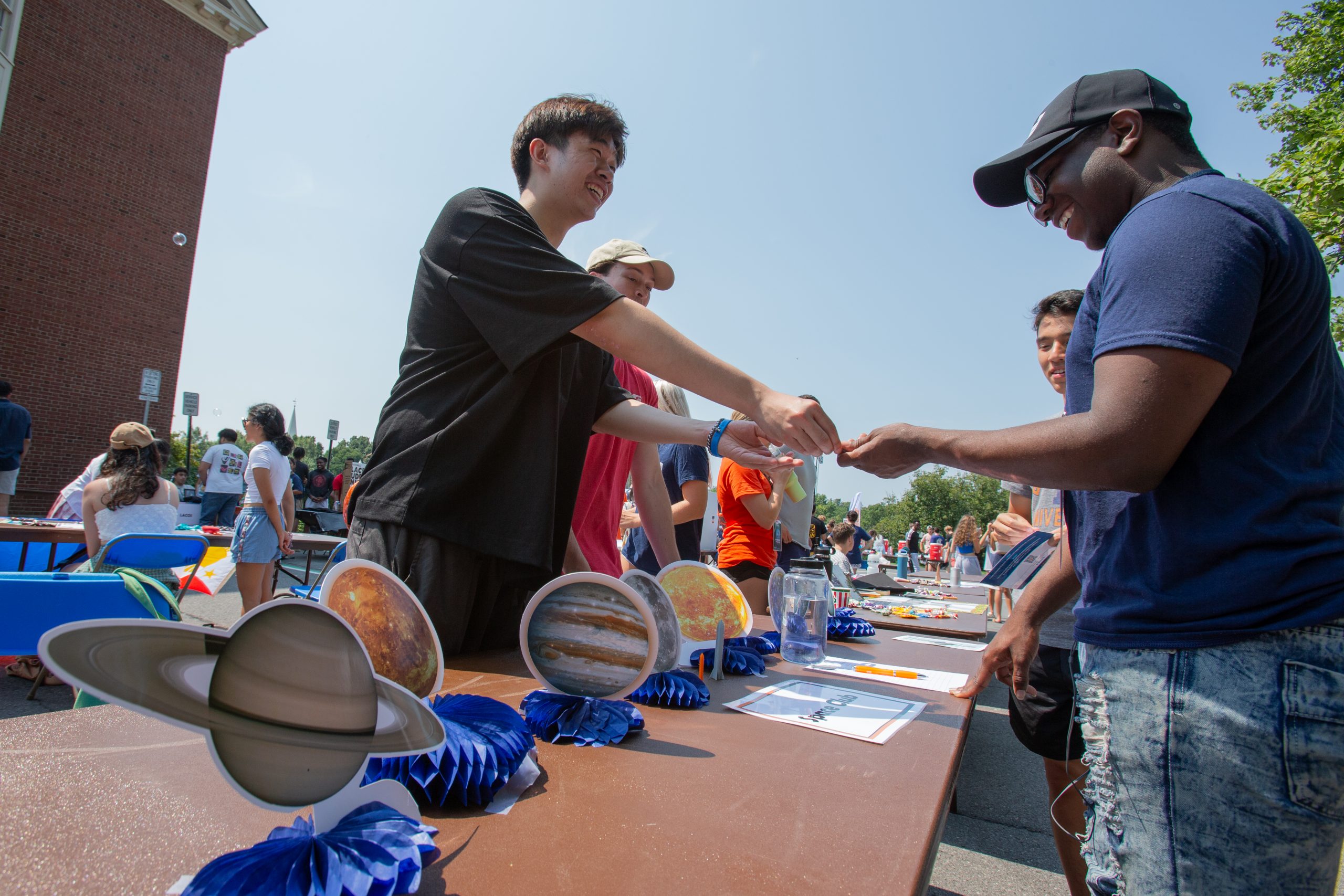 A male student passes a piece of paper to another male student over a table outdoors at the Activities Unlimited fair. There are paper cut outs of planets on the table.